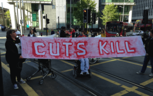 disabled people holding sign in middle of road "cuts kill". Wheelchairs and other walking aids are visible