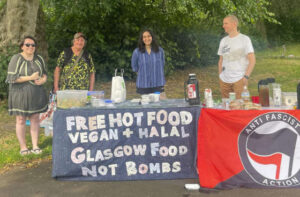 Four people standing behind a stall offering free food. One banner: "free hot food vegan + halal Glasgow food not bombs" The other: "Antifascist action"